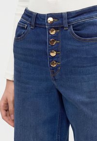 Μπλε τζιν από denim με πέντε ορατά χρυσά κουμπιά και λευκό ριμπ μπλουζάκι με μακριά μανίκια μερικώς ορατό.