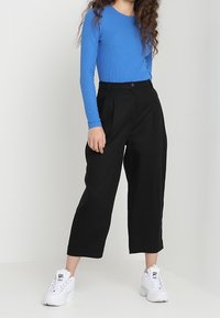 Donna che indossa un top a maniche lunghe di un brillante blu, pantaloni cropped neri a vita alta e larghi, e sneakers bianche spesse, in piedi davanti a uno sfondo uniforme.