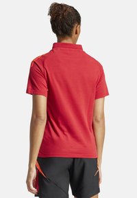 Rotes Poloshirt mit Kragen und kurzen Ärmeln, das orangefarbene Akzente an den Schultern aufweist. Wird mit schwarzen Sportshorts getragen.