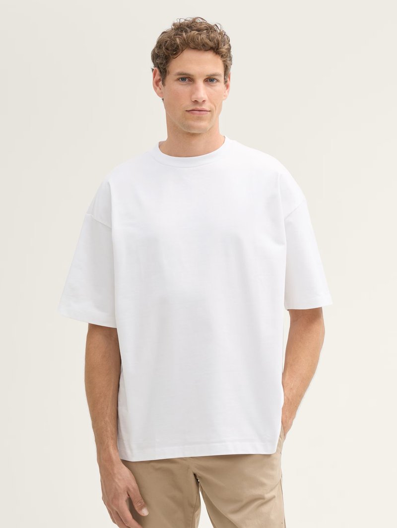 Fehér, túlnyomórészt pamut oversized póló, rövid ujjak, kerek nyakkivágás és sima textúra jellemzi, bézs nadrággal viselve.