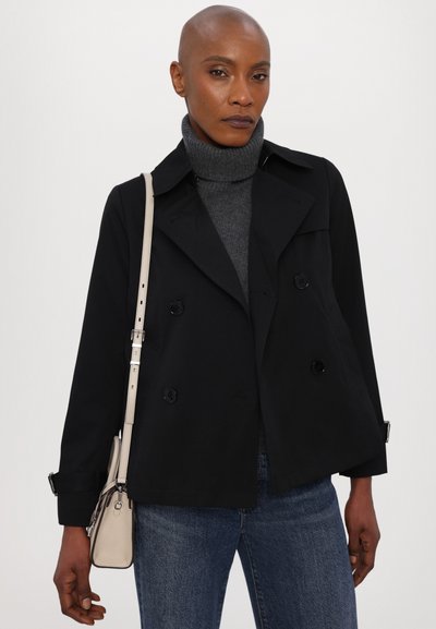 Veste noire croisée avec col, manches longues et accents de boutons. Associée à un pull à col roulé gris et un jean bleu, tenant un petit sac.