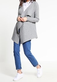 Harmaa neulottu neuletakki, jossa on avoin etuosa, laskeutuva malli ja pitkät hihat, yhdistetty sinisiin farkkuihin ja valkoisiin tennareihin.