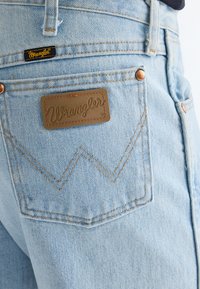 Világoskék farmer hátul zseb, barna Wrangler bőrpárnával és varrott "W" mintával, vörösréz szegecsekkel és apró fekete Wrangler címkével.
