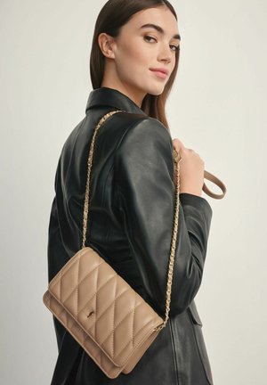 Kobieta w czarnej skórzanej kurtce niesie beżową pikowaną torebkę na ramię ze złotym łańcuszkiem na tle jednolitego tła.