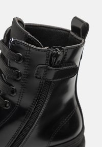 Botte cheville en vernis noir avec un col texturé, un devant lacé, une fermeture éclair sur le côté et une semelle robuste avec des détails de couture.