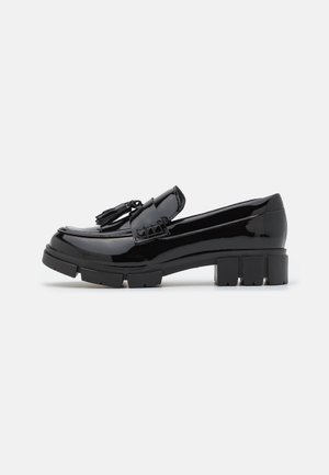 Mocassin en cuir verni noir avec semelle épaisse et détail de pompon sur le dessus, présenté en profil latéral sur un fond blanc.