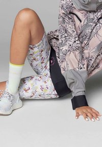Pantaloni sportivi a fantasia floreale in grigio chiaro e bianco, abbinati a una giacca grigia a motivi multipli. Calzini sportivi verde chiaro e sneakers grigie.