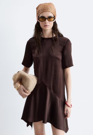 Femme portant une robe marron foncé à manches courtes, un foulard orange à motifs, des lunettes de soleil ambre, tenant un sac en tricot beige avec des bracelets dorés à un bras.