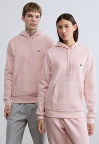 Ροζ φούτερ με κουκούλα σε ανοιχτή απόχρωση, τσέπες καγκουρό, μανσέτες και τελειώματα με ριμπ ύφανση, στολισμένα με το πράσινο λογότυπο Lacoste. Γκρι παντελόνια φόρμας συμπληρώνουν τα φούτερ.