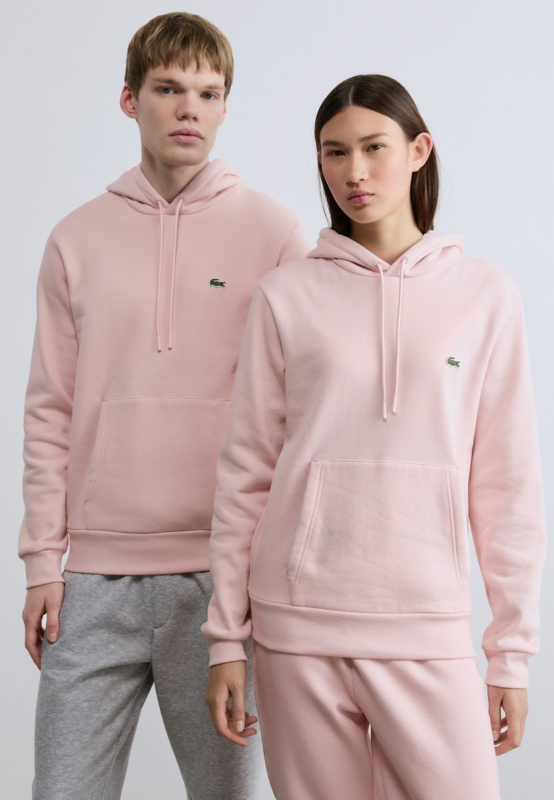 Ροζ φούτερ με κουκούλα σε ανοιχτή απόχρωση, τσέπες καγκουρό, μανσέτες και τελειώματα με ριμπ ύφανση, στολισμένα με το πράσινο λογότυπο Lacoste. Γκρι παντελόνια φόρμας συμπληρώνουν τα φούτερ.