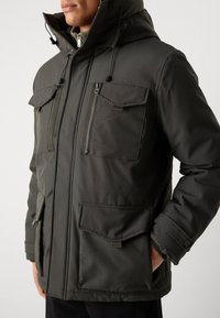 Giacca a vento verde scuro con cappuccio, presenta una chiusura con zip, due tasche anteriori con pattina e una tasca con zip aggiuntiva. Realizzata in tessuto testurizzato con dettagli regolabili.