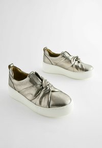 Metallic silver slip-on shoes with a knotted design, white platform sole, and textured finish. Features a pull tab at the back.