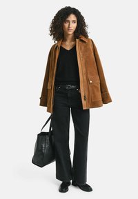 Braune Wildlederjacke mit großen Vorderaschen, kombiniert mit einem schwarzen V-Ausschnitt-Pullover und hochgeschnittenen schwarzen Jeans. Schwarze Tote-Tasche und Schuhe.