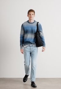 Ανοιχτό μπλε και γκρι γκράντ ζακέτα, ανοιχτό τζιν και μαύρες παπούτσια. Άντρας κρατάει μια μαύρη τσάντα, στέκεται μπροστά σε ουδέτερο φόντο.