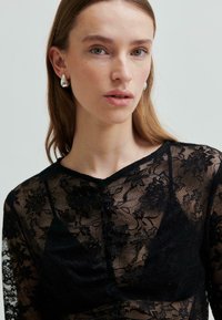 Młoda kobieta o jasnobrązowych włosach, nosząca srebrne kolczyki w kształcie łezki oraz czarną koronkową bluzkę w kwiaty, na jednolitym tle.