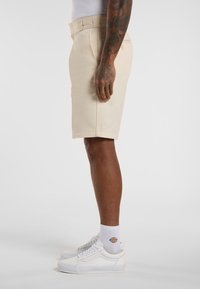 Kniehohe khakifarbene Shorts mit flachem Bund und Gürtelschlaufen, kombiniert mit weißen Sneakers und gemusterten Socken, die eine taillierte Silhouette zeigen.