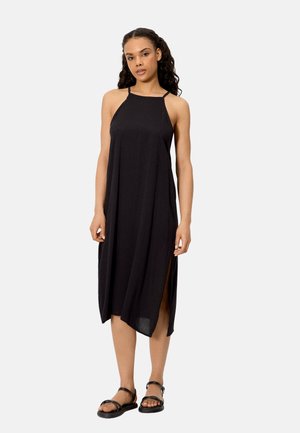 Mujer con cabello largo y rizado, vestida con un vestido midi negro sin mangas con una abertura lateral y sandalias planas negras con tiras, de pie frente a un fondo liso.