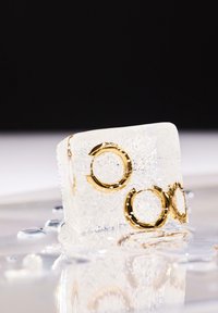 Cubo di ghiaccio trasparente con anelli d'oro incorporati, caratterizzato da una superficie liscia e bolle. Gocce d'acqua circostanti riflettono la luce su una superficie.