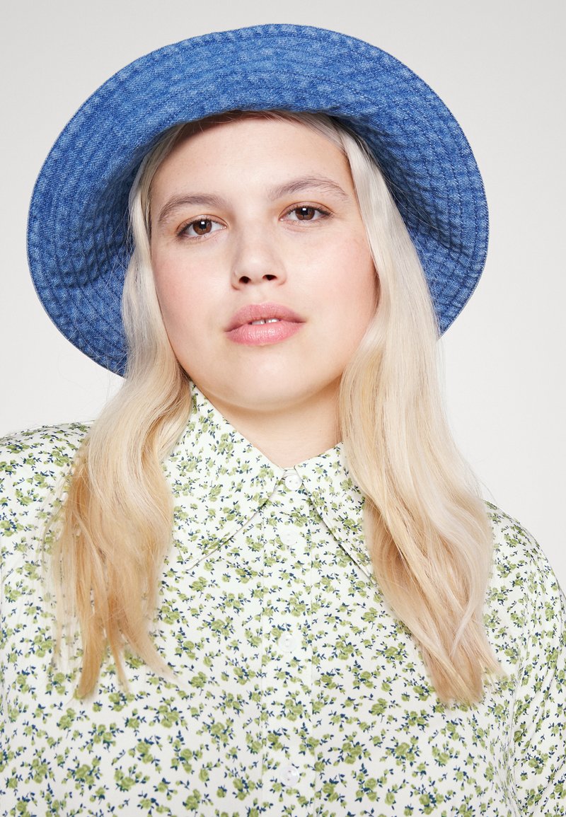 Cappello a secchiello blu con una finitura texturizzata, abbinato a una camicia bianca con bottoni che presenta un motivo floreale verde e blu.
