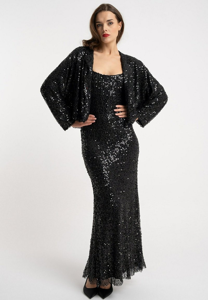 Czarna wieczorowa suknia wykonana z cekinowej tkaniny, o dopasowanym kroju, z dopasowaną kurtką z szerokimi rękawami. Podkreślona głębokim dekoltem.