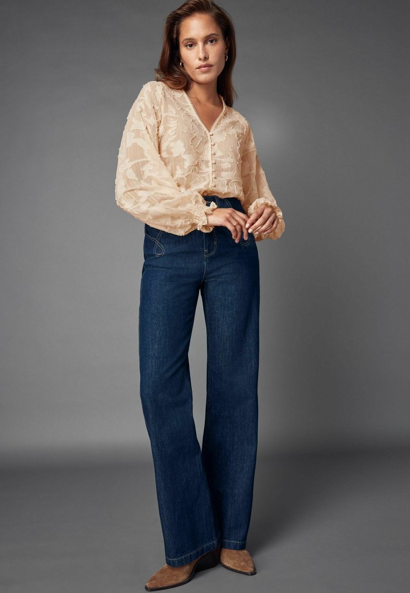 Blouse beige transparente avec motif floral texturé et fermeture à boutons, associée à un pantalon en denim foncé taille haute évasé et des chaussures à talons marron.