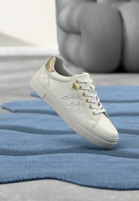 Baskets blanches avec accents dorés et motif en relief, suspendues en l'air au-dessus d'un tapis bleu texturé sur un sol gris.