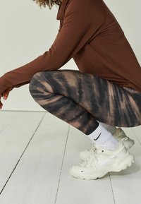 Ruskea pitkähihainen paita, mustat ja ruskeat kravattivärjätyt legginsit, valkoiset Nike-tennarit ja valkoiset sukat henkilöllä, joka on kyykyssä valkoisella puulattialla.