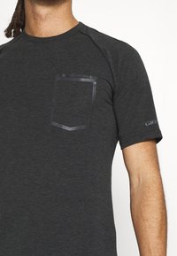 Kortärmad sporttröja i mörkgrått tyg med en texturerad yta, med en bröstficka och en logotyp på ärmen.