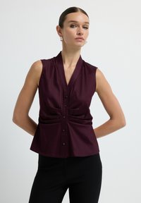 Blouse sans manches bourgogne avec un design enroulé et des boutons à l'avant. Présente de légères plis sur les côtés, fabriquée en tissu doux.
