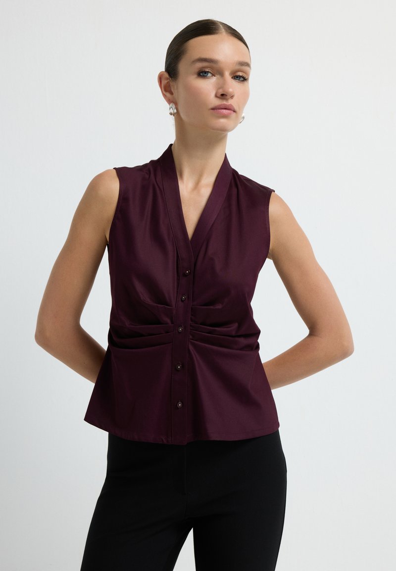 Blouse sans manches bourgogne avec un design enroulé et des boutons à l'avant. Présente de légères plis sur les côtés, fabriquée en tissu doux.