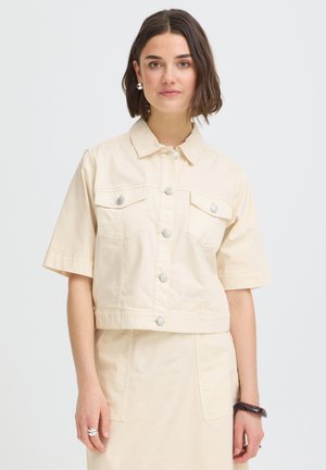 Femme aux cheveux bruns mi-longs, portant une veste crème à manches courtes boutonnée et une jupe assortie, accessoirisée de boucles d'oreilles, d'une bague et d'un bracelet.