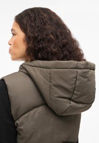 Maslinasto zelena prsluk jakne na jastučiće s velikim kapuljačom, prošivenim dizajnom i glatkom teksturom, nošen preko crne majice dugih rukava.