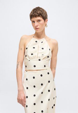 Vrouw met kort haar die een crèmekleurige haltertop en rok met zwarte stippen draagt, staand tegen een effen lichte achtergrond.