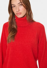 Raudonas megztinis su aukšta apykakle, pagamintas iš minkštos tekstūros, su laisvu siluetu ir raukšlėta apykakle, parodantis šiek tiek margą raštą.