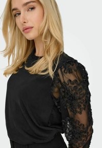 Top nero con maniche lunghe trasparenti decorate da appliqué floreali, realizzato in tessuto morbido e con una vestibilità rilassata. Texture liscia, design elegante.