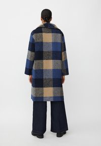 Ternet frakke i nuancer af blå, mørk marineblå og beige. Blød tekstur med en bred krave og lange ærmer. Denim bukser og kraftige sorte støvler.