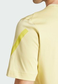 Světle žlutá košile s krátkým rukávem, hladkou texturou a výrazným žlutým pruhem na rameni, s kulatým výstřihem a detaily v šití.