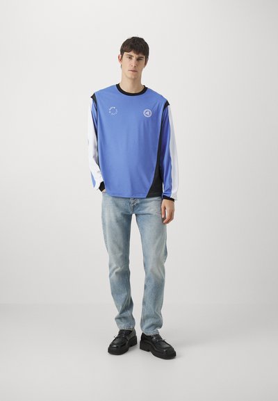 Blue sweatshirt with black and white accents, logo on chest, paired with light blue jeans and black shoes. Smooth fabric texture.