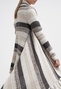 Mujer con un suéter largo de punto suelto con amplias rayas horizontales en beige, marrón y gris oscuro, sujetando el dobladillo que fluye.