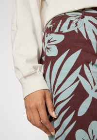 Main reposant contre la hanche portant un pantalon à motifs floraux bleu et bordeaux et un haut beige à manches longues avec poignets froncés.