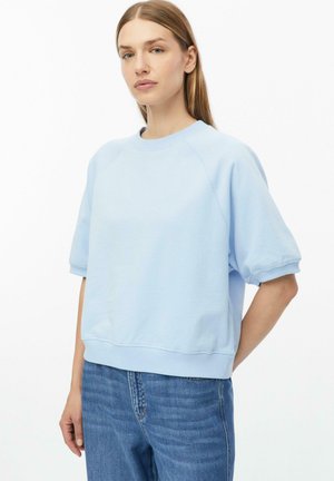 Femme adulte aux cheveux raides portant un sweat-shirt bleu clair à manches courtes et un jean bleu, debout avec une main derrière le dos.