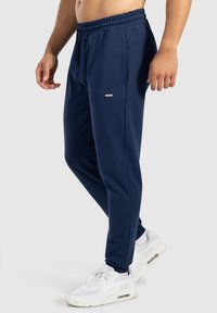 Námořnické jogger kalhoty vyrobené z měkké tkaniny, s elastickým pasem, kapsami a malým brandingovým prvkem poblíž kyčle. Sestavené s bílými teniskami.