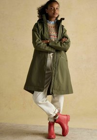 Cămașă impermeabilă de culoare verde măsliniu cu glugă, pulover cu model, pantaloni albi și cizme roșii din cauciuc. Textură netedă și croială lejeră.