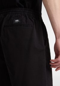 Pantaloni corti leggeri neri con cintura elastica, tasche laterali e una tasca sul retro con un'etichetta logo Vans. Texture liscia.