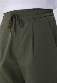 Olivgröna byxor med elastisk midja, dragsko, och sidofickor. Tillverkade av mjukt, strukturerat tyg med veck.