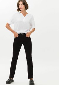Weiße Bluse mit kurzen Ärmeln und Kevinskragen, kombiniert mit schwarzen, schmal geschnittenen Jeans. Schwarze, flache Schuhe runden das Outfit ab.