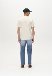 Világosbézs rövid ujjú póló, fakó kék farmernadrág és barna cipő. A személy zöld sapkát visel, és láthatók a fülhallgatói.