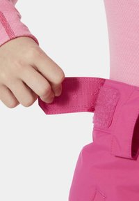 Růžový textilní pásek s uzavíráním na suchý zip držený v ruce. Pásek ladí s růžovým oblečením, má hladký povrch a strukturovaný tvar.