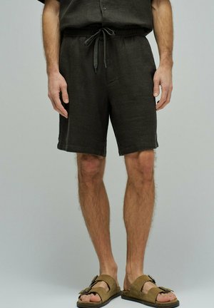 Pantalones cortos de hombre de color gris oscuro con cordón, usados con sandalias marrones con hebilla, mostrados desde la cintura hasta los pies sobre un fondo liso.