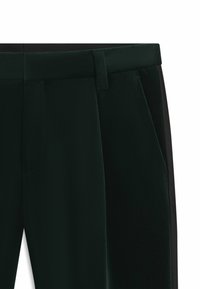 Dunkelgrüne Hose mit glatter Textur, seitlichen Taschen und einem schwarzen Einsatz entlang der äußeren Naht. Taillierte Passform mit flachem Bund.